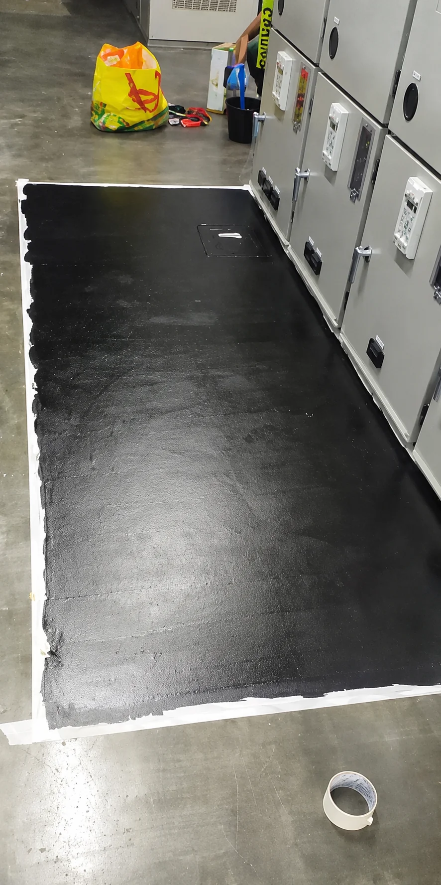 Black conductive base coat applied to a taped-off strip of floor running the length of a switchgear aisle, with a floor access hatch left visible.