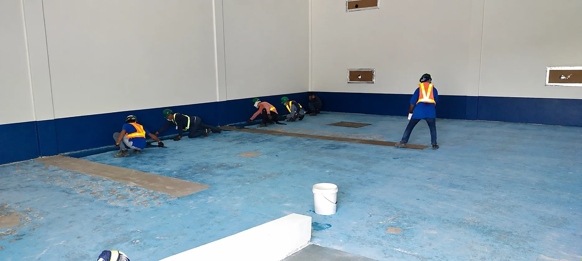 A crew in hard hats and hi-vis vests kneeling along the perimeter of a room, preparing the edge of a worn blue floor coating before recoating.
