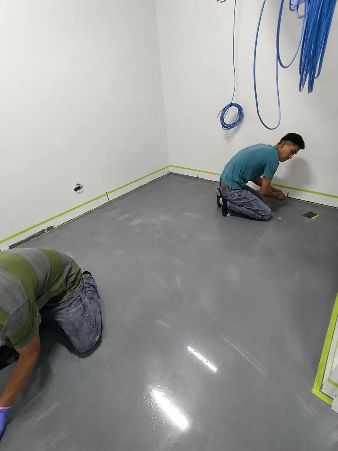 Two installers kneeling to detail the edges of a grey ESD epoxy topcoat against masking tape along the wall.