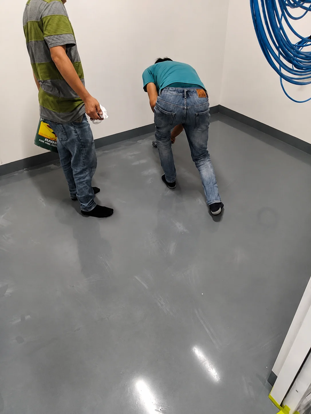 Two installers working on a freshly poured grey epoxy floor in a small equipment room with blue cabling coiled on the wall.