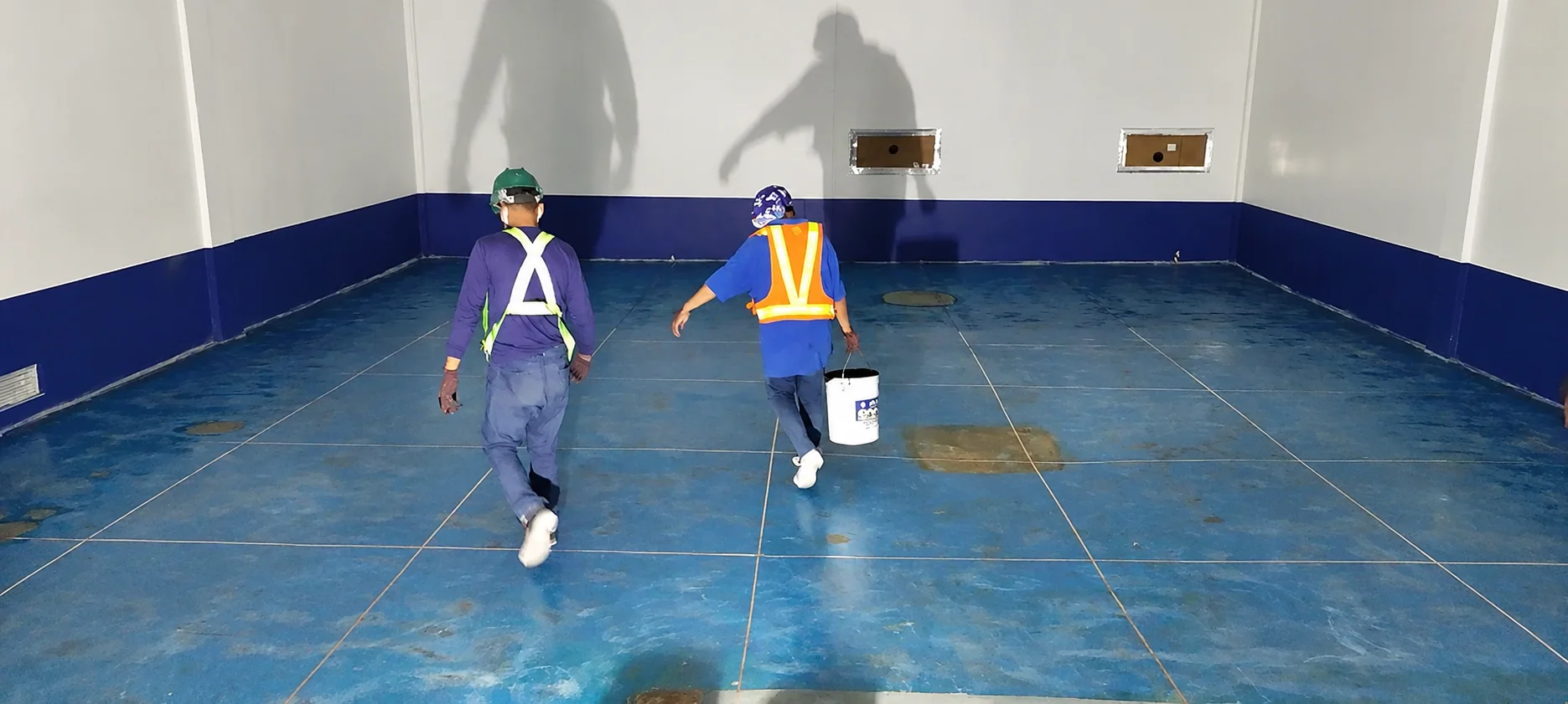 Two workers carrying a paint pail across an existing blue ESD tile floor with visible grid joints and stained patches, before replacement.