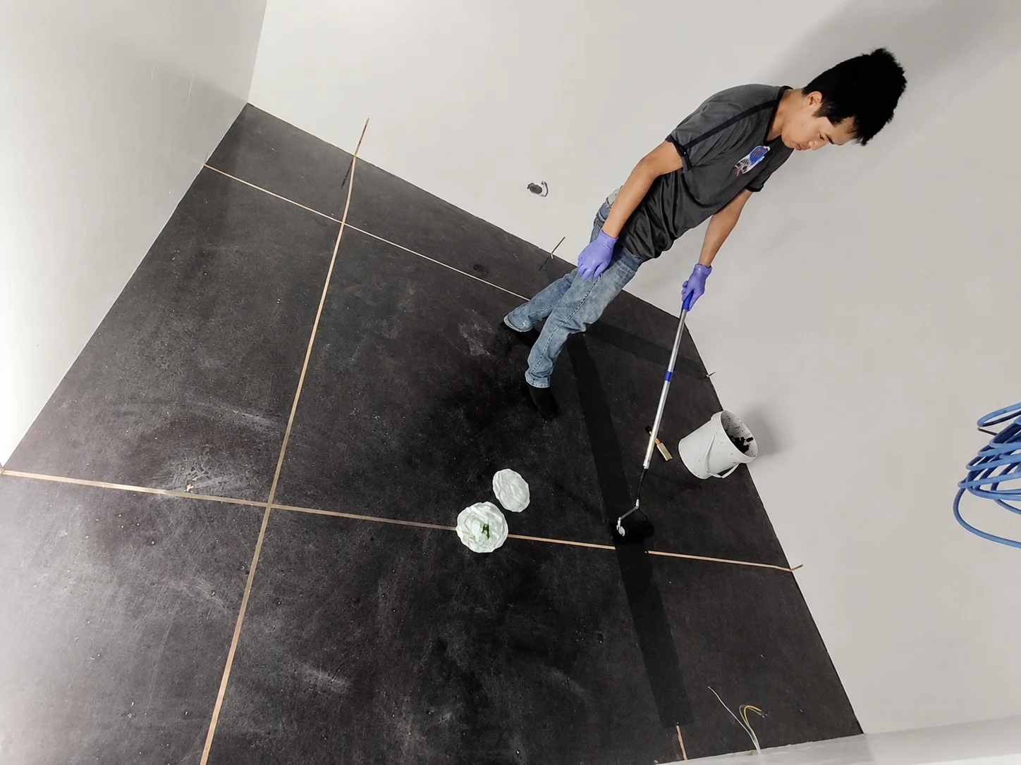 A technician in gloves using a long-handled roller to apply a black conductive coating over a floor with a copper grounding grid.